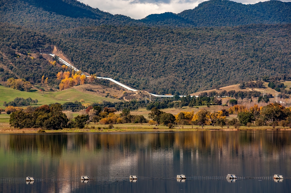 Scenic view of a water pipeline running down a hillside with autumn trees reflecting in the calm waters of a reservoir.