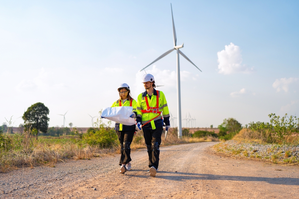 Two engineers with safety vests and helmets reviewing plans in front of a large wind turbine in a field, symbolising renewable energy infrastructure.
